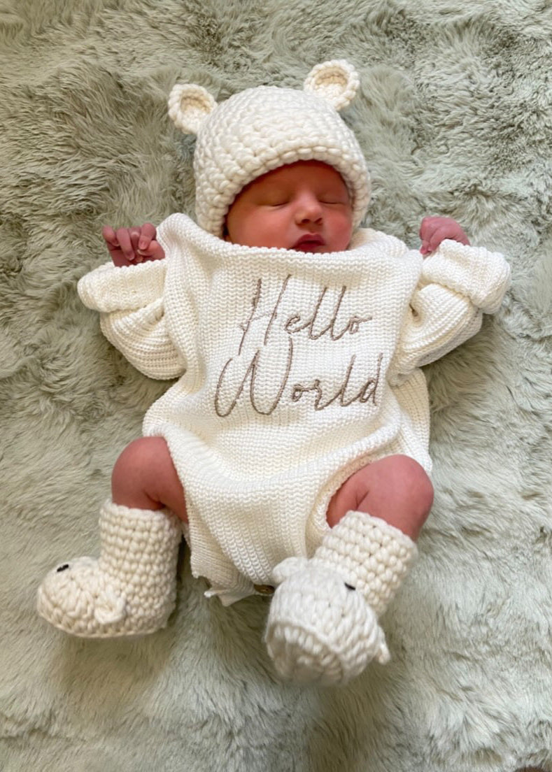 Newborn baby wearing a white knitted outfit with 'Hello World' text, lying on a fluffy surface.
