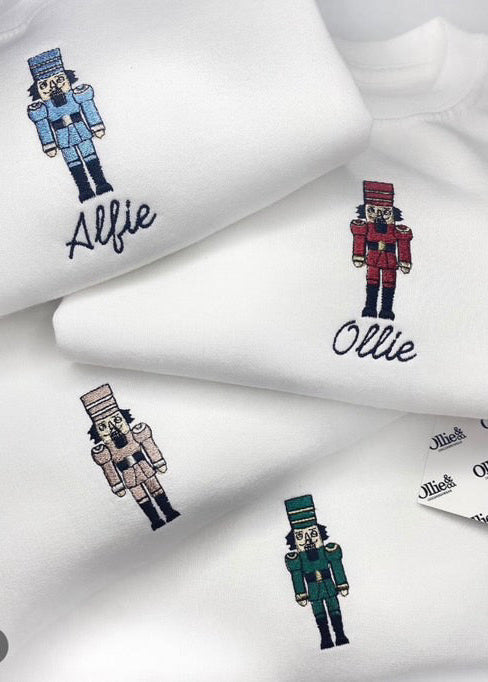 White sweaters with nutcracker character designs on the front, personalized with the names 'Alfie' and 'Ollie' on the sleeves.
