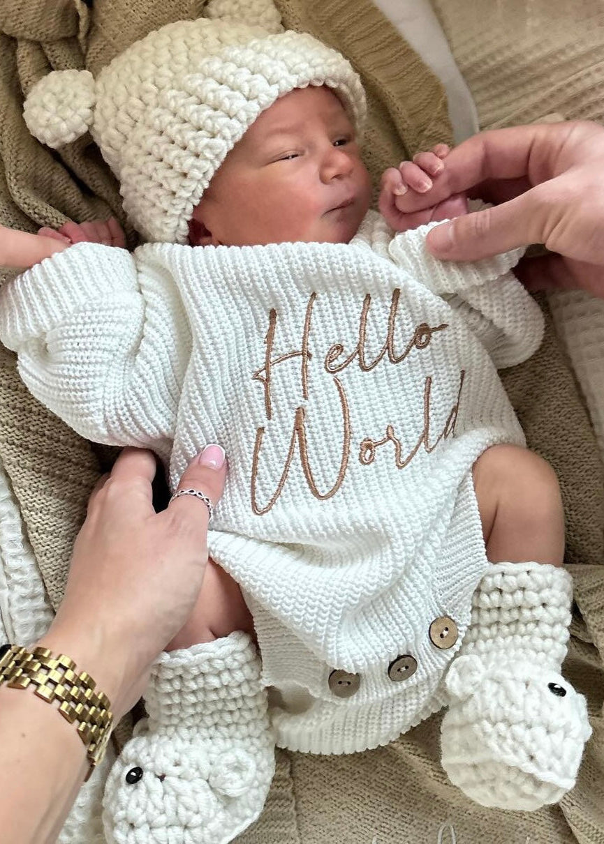 Newborn baby in a 'Hello World' outfit being held by two people, surrounded by beige blankets.