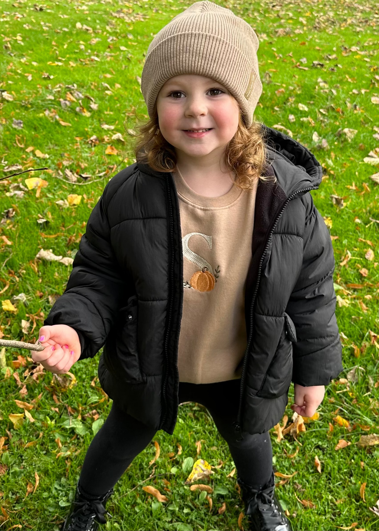 Child wearing a black puffer jacket and beige beanie standing on grass with scattered leaves.