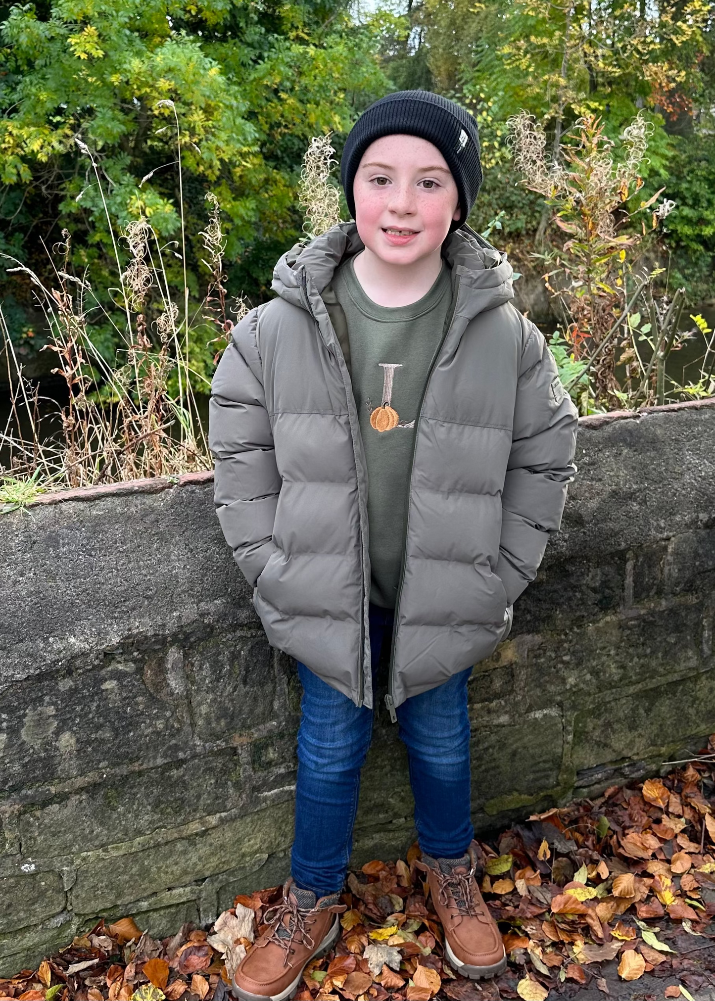 Child wearing a gray puffer jacket and black beanie standing against a stone wall with autumn leaves and foliage.