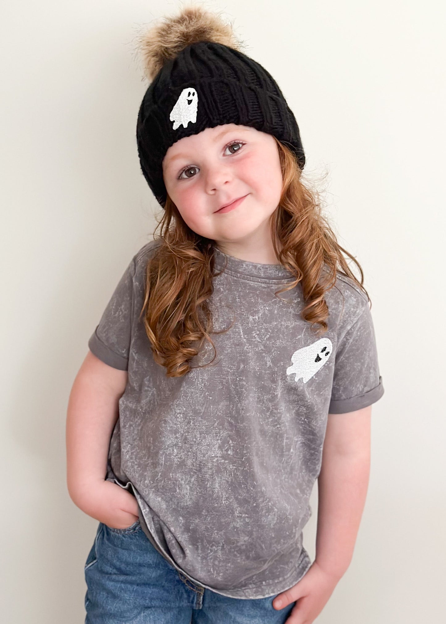 Child wearing a black knit hat with a ghost design and a matching gray t-shirt with a ghost design on a plain background.