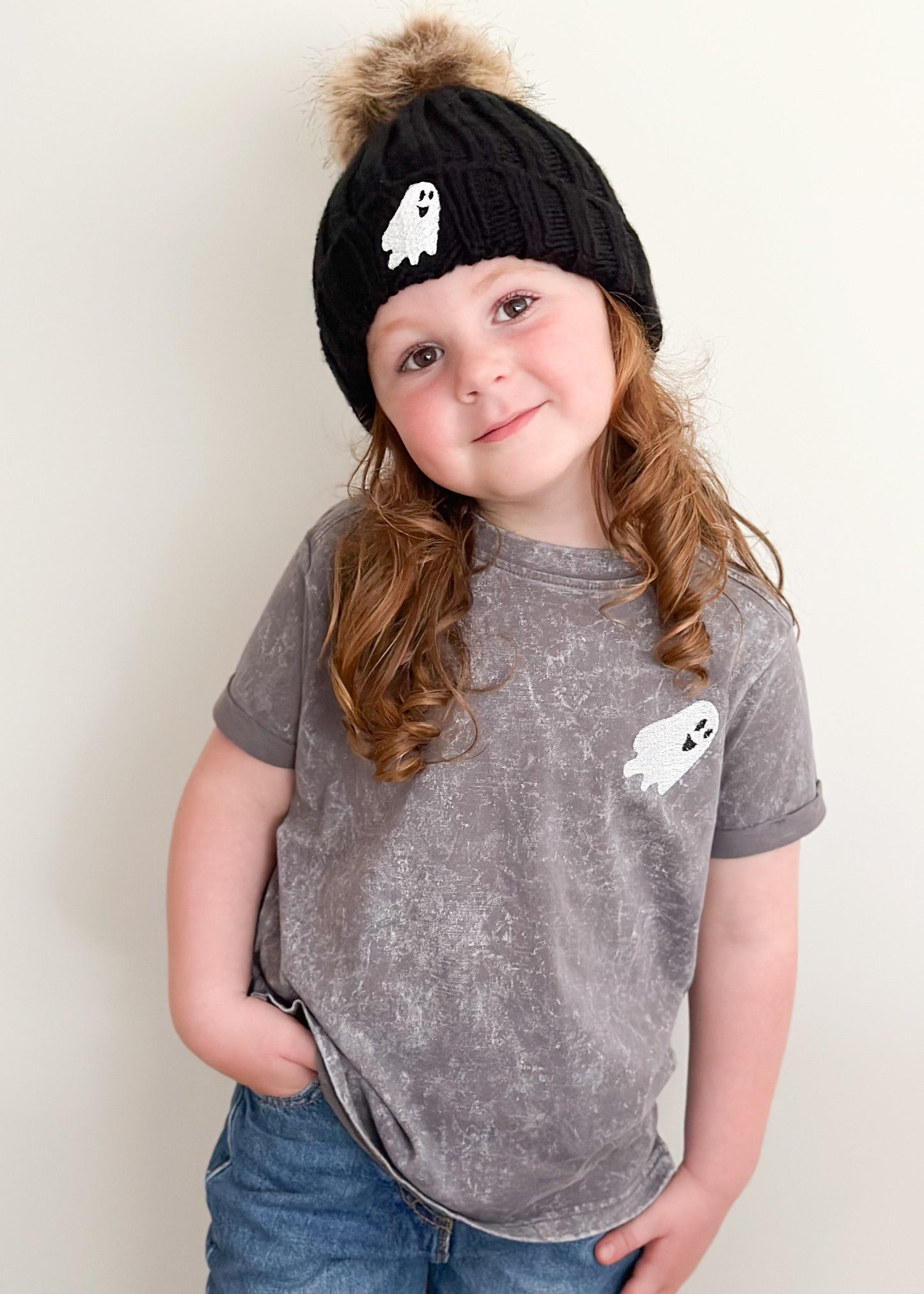 Child wearing a black knit hat with a ghost design and a matching gray t-shirt with a ghost design on a plain background.