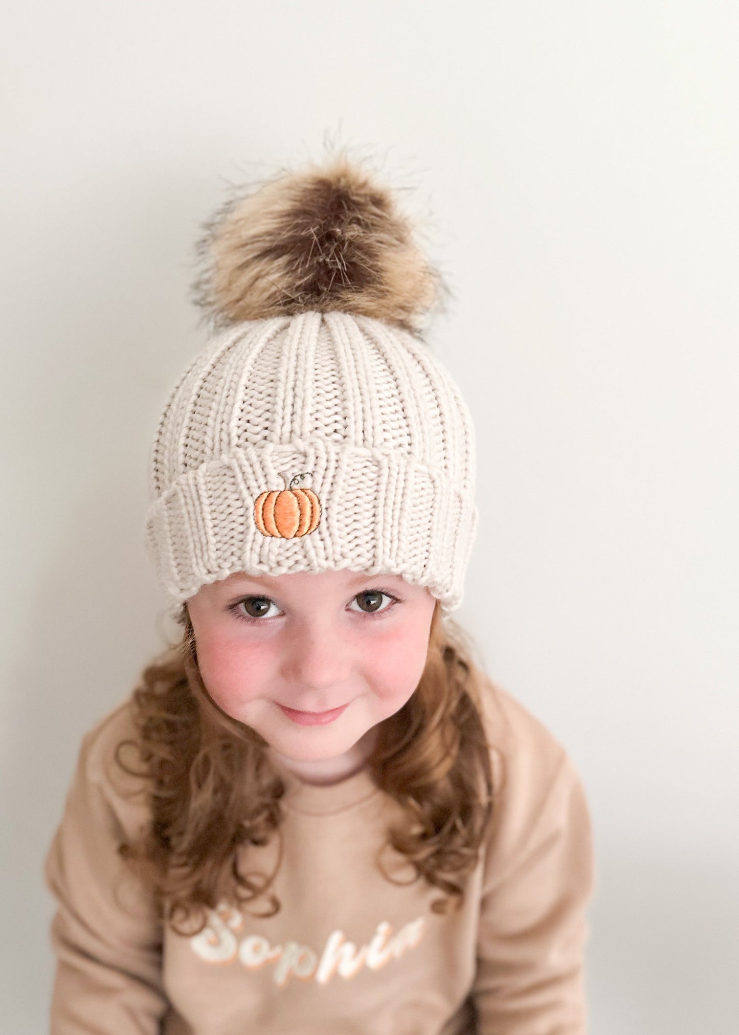 Child wearing a cream knit beanie with a pom-pom and pumpkin charm, sitting against a plain background.