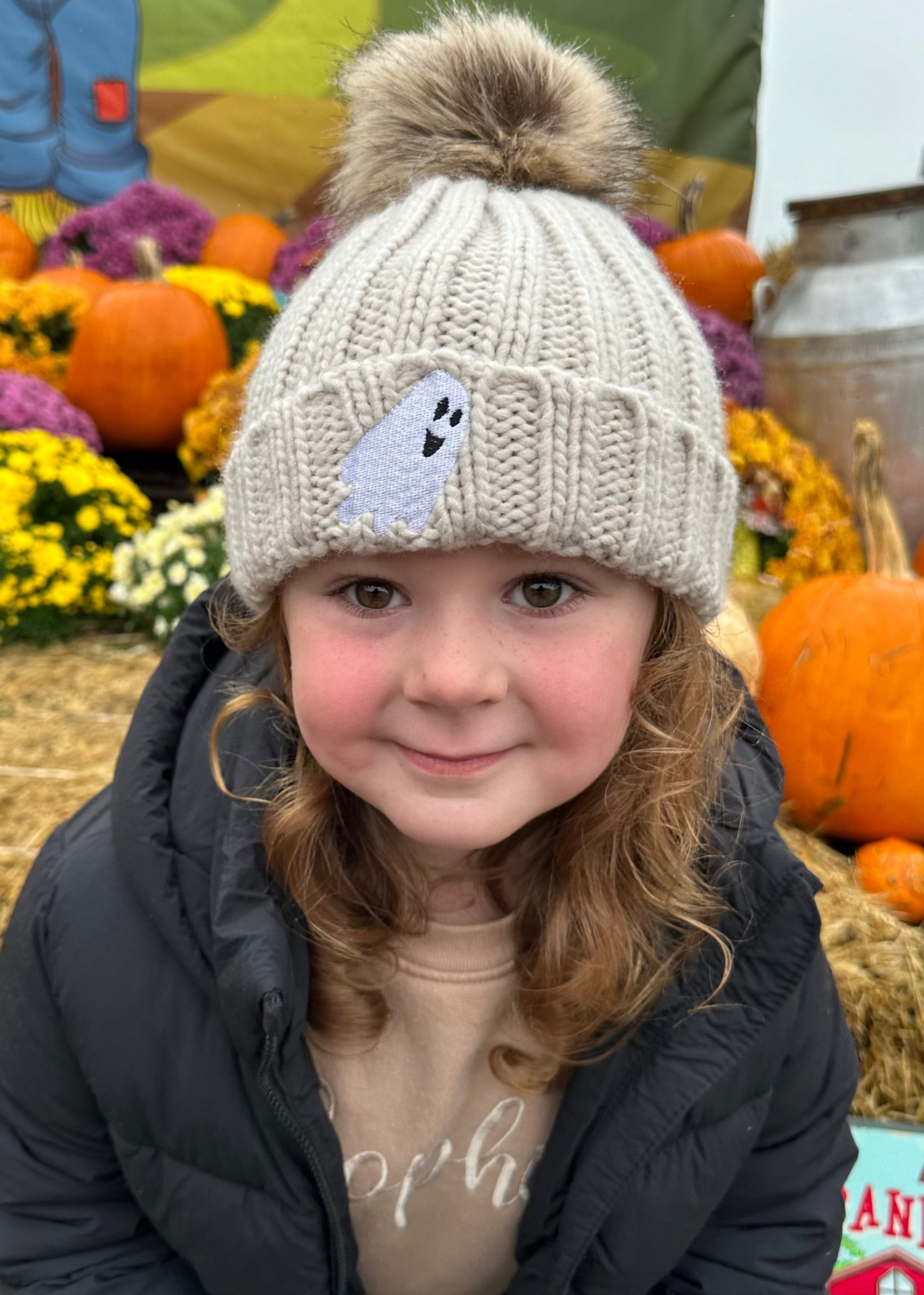 Child wearing a knit hat with a ghost design in an autumn setting with pumpkins and flowers.