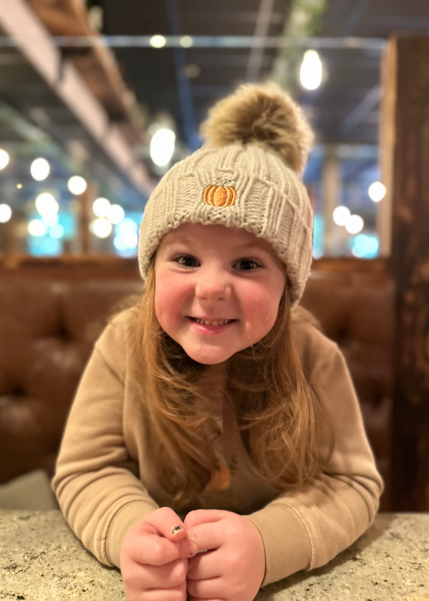 Child wearing a beige knit hat with a pumpkin design, sitting indoors.