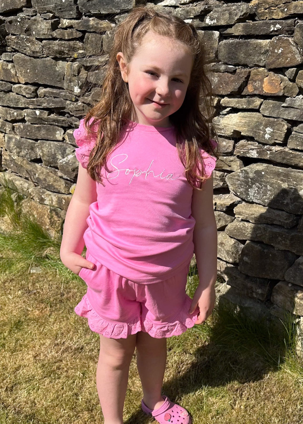 Young girl in a pink outfit standing in front of a stone wall.