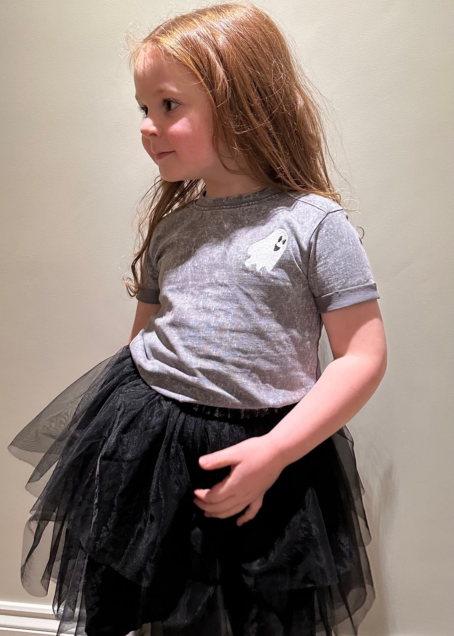 Young girl wearing a gray t-shirt and black tutu standing against a plain wall.