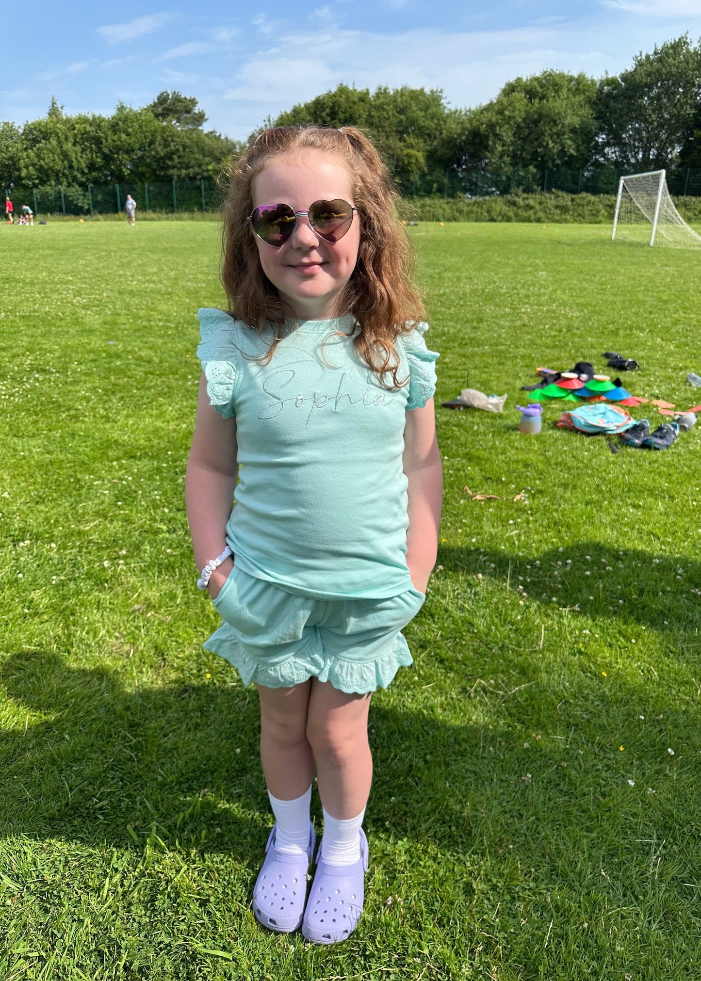 Young girl in a light blue outfit standing on a grassy field with playground equipment in the background.