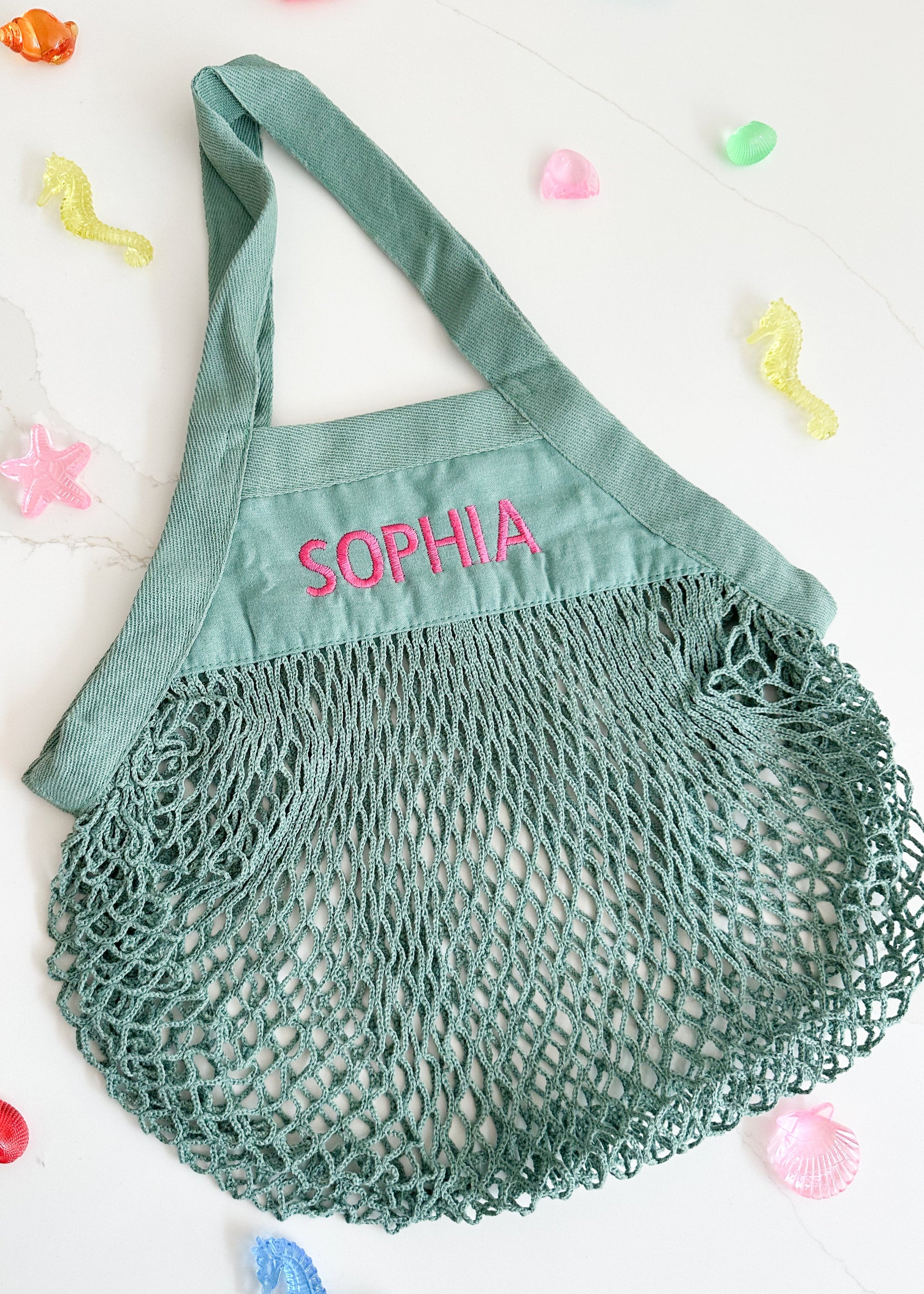 Green mesh bag with 'SOPHIA' embroidered on it, surrounded by colorful candy on a white background.