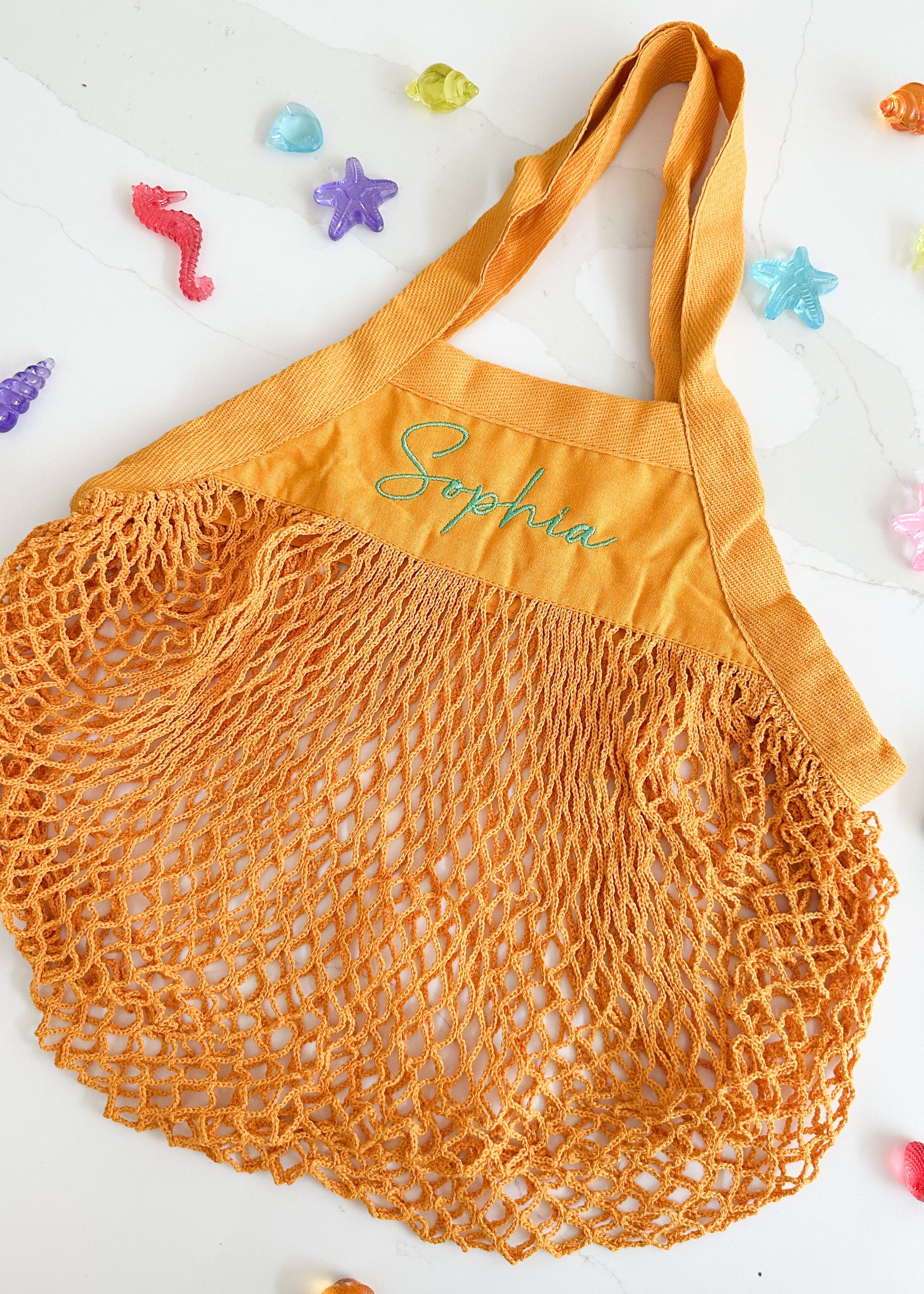Orange mesh shopping bag with 'Sophie' embroidered on it, on a white background with colorful star-shaped objects.