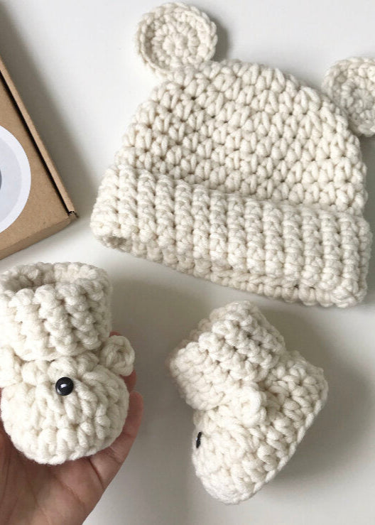 White knitted baby hat and booties set with a smiley face box on a light background