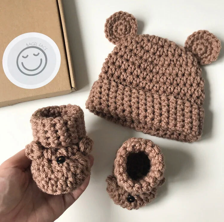 Knitted brown baby hat and booties set on a white surface with a cardboard box in the background.