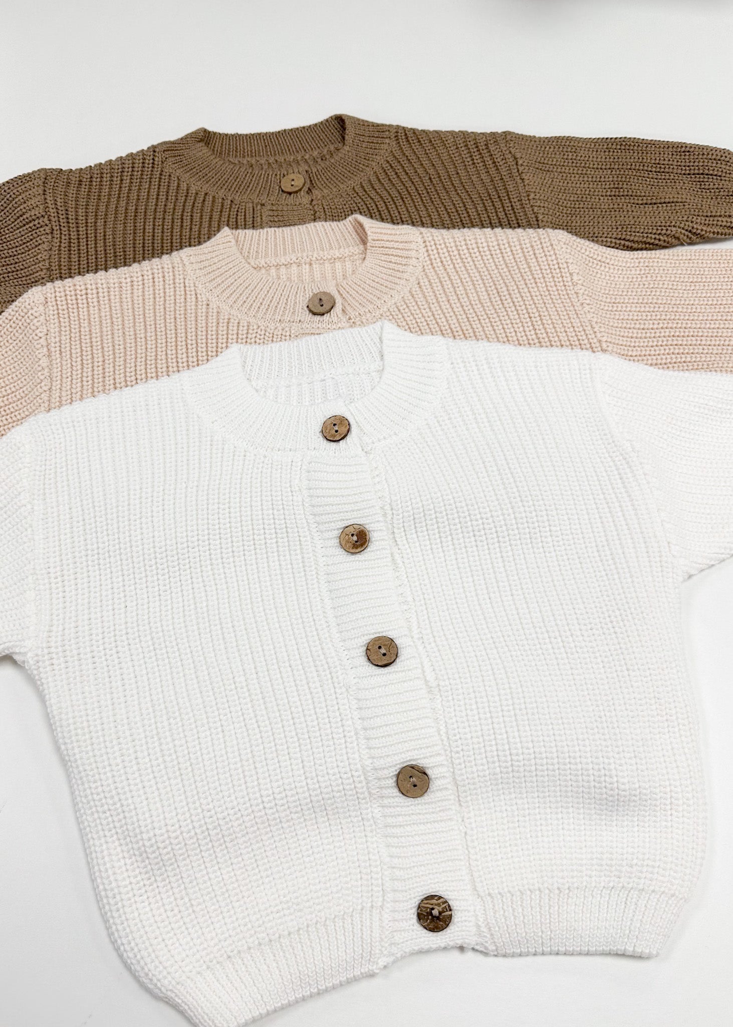 Three knit cardigans in brown, beige, and white with button details on a light gray background.