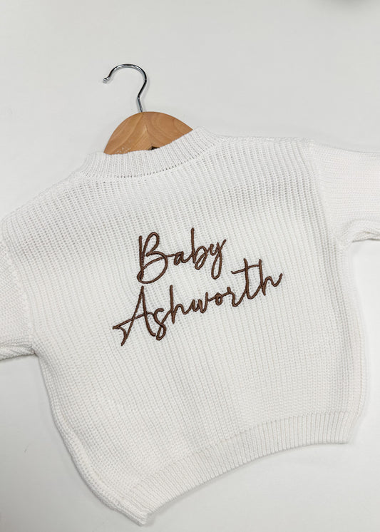 White sweater with 'Baby Ashworth' text on a hanger against a white background