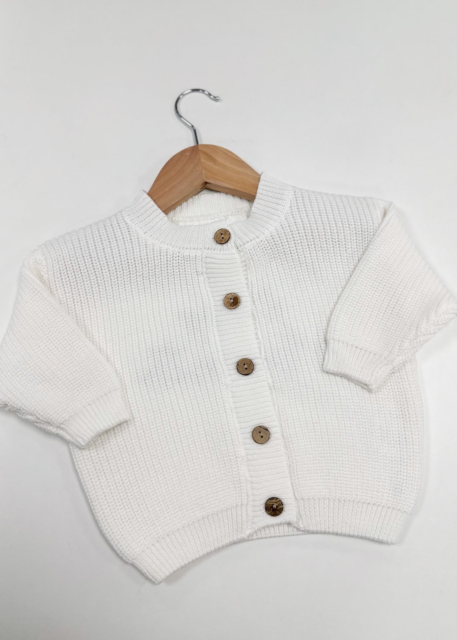White knitted cardigan with brown buttons on a wooden hanger against a light gray background