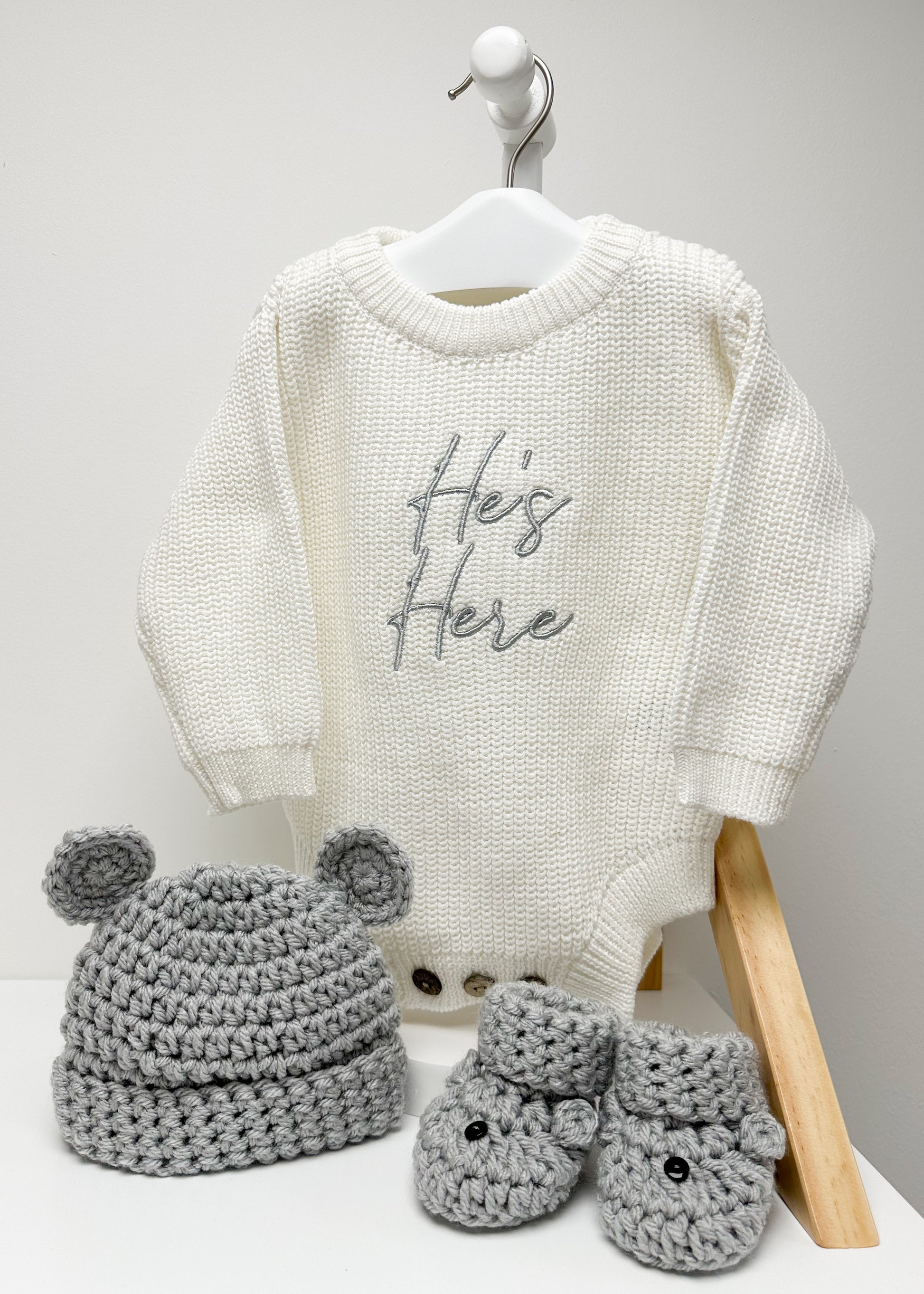 White baby sweater with 'He's Here' text, gray crocheted hat, and booties on a white background