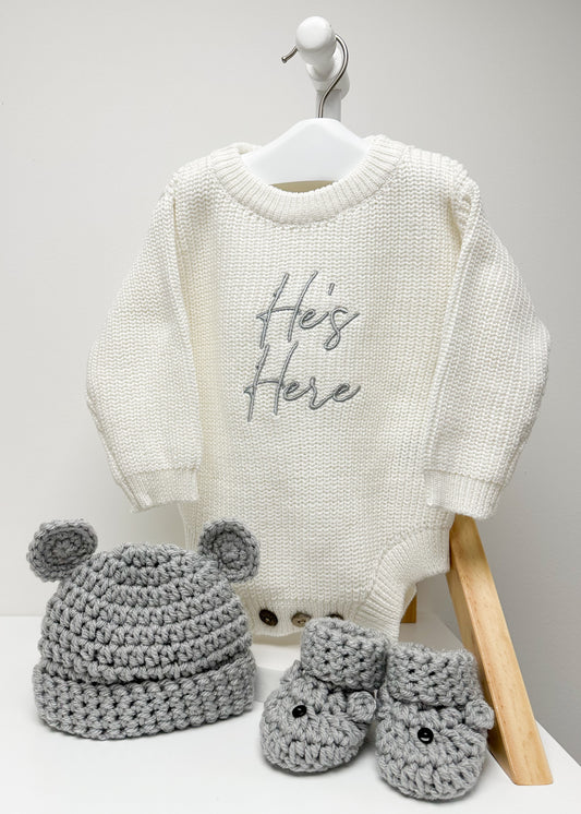 White baby sweater with 'He's Here' text, gray crocheted hat, and booties on a white background