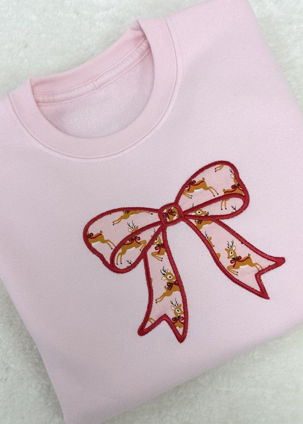 A pink fleece crew neck sweatshirt with a red embroidered Christmas bow appliqué on the front.