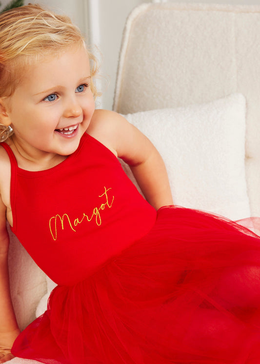 Young girl in a red dress with 'Margot' embroidered on it, sitting on a white couch.