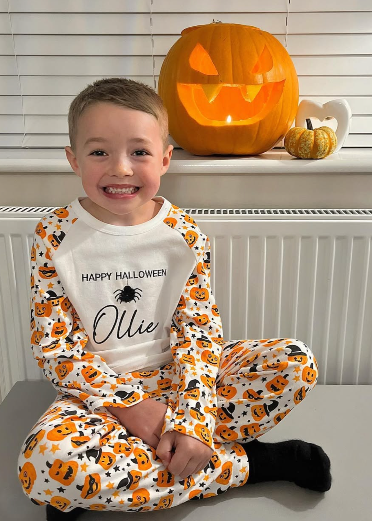 Child wearing Halloween-themed pajamas with a pumpkin design, sitting in front of a carved pumpkin.
