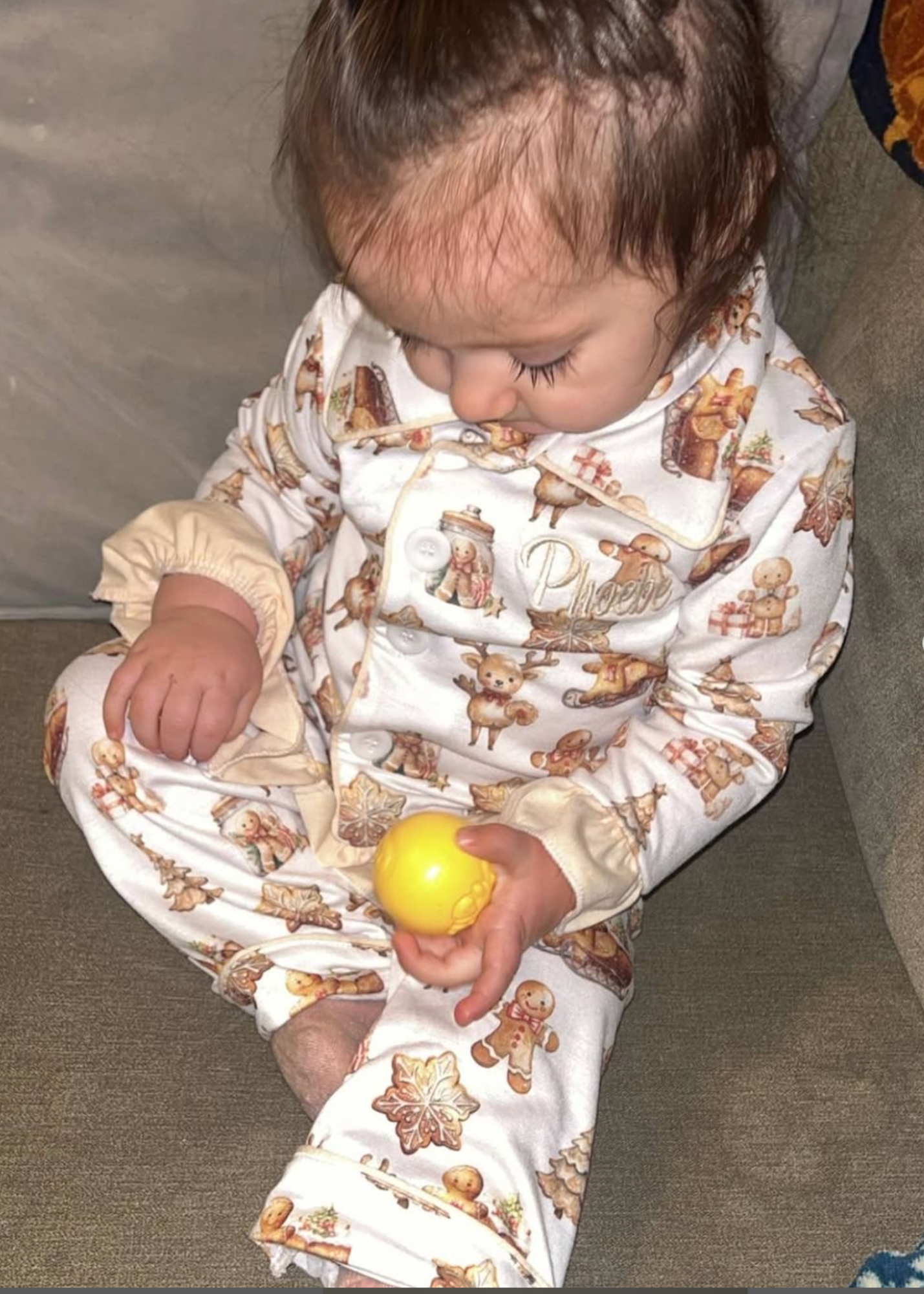 Baby wearing a patterned outfit holding a yellow ball on a carpeted floor.