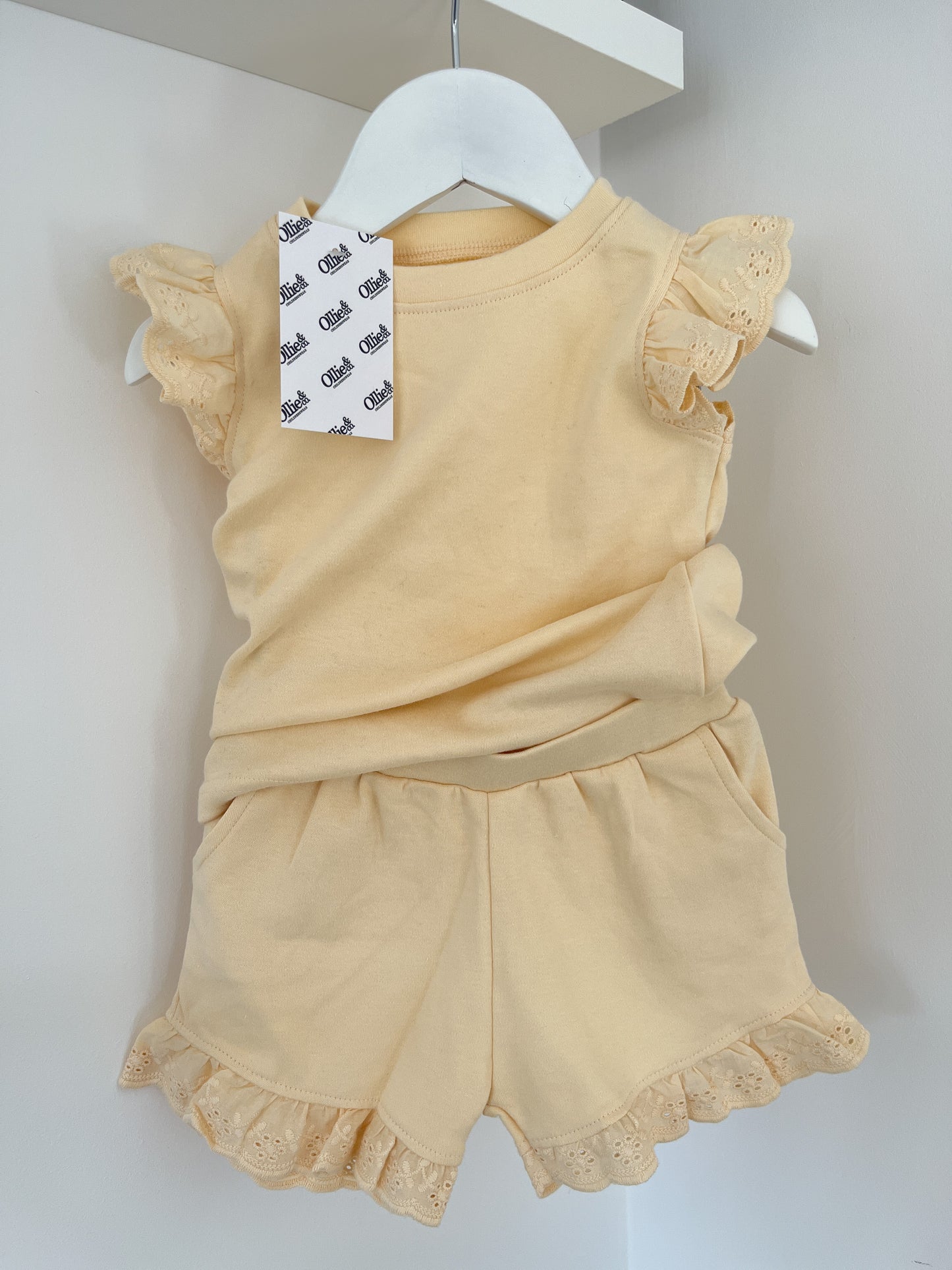 A selection of children's frill tee and shorts sets hanging on hangers, with colors including blue, pink, yellow, and brown.
