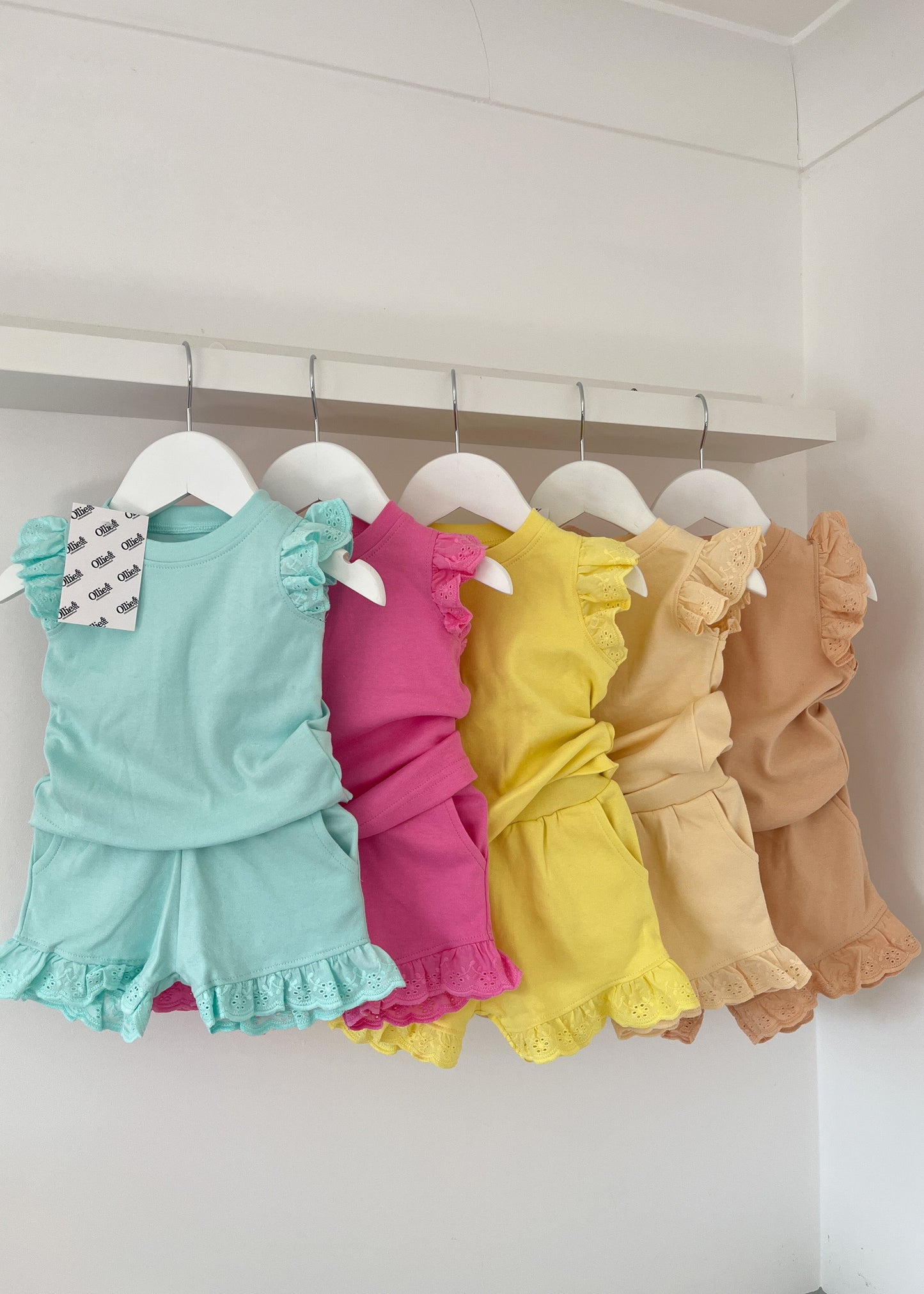 A selection of children's frill tee and shorts sets hanging on hangers, with colors including blue, pink, yellow, and brown.