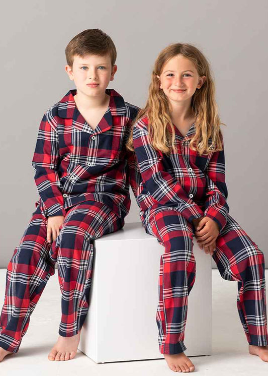 Two children, a boy and a girl, sitting on a white surface wearing red and navy tartan Christmas pyjamas with small left chest embroidery.