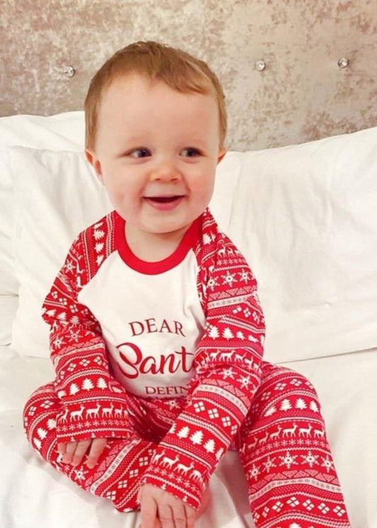 A baby sitting on a bed wearing red and white Christmas themed pyjamas with a 'Dear Santa' text on the chest and a Santa Claus design.