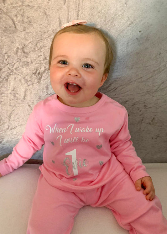 A smiling baby wearing pink birthday pyjamas with text on the shirt.