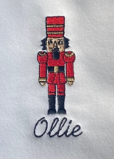 A embroidered nutcracker romper with the name 'Ollie' on the front, featuring a character in a red uniform with black details and a grey background.
