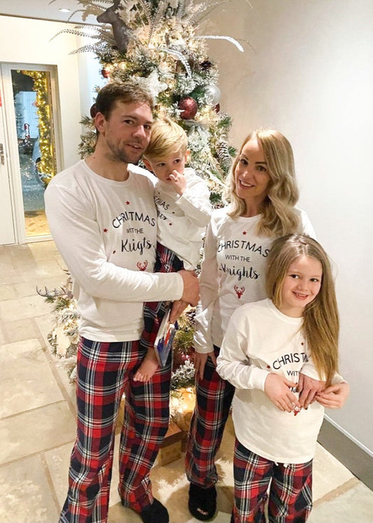 A family of five, including two adults and three children, standing in front of a decorated Christmas tree and festive wreaths. All members are wearing matching red and navy tartan Christmas pyjamas with a 'Christmas Knight' text on the adults' shirts.