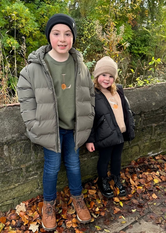 Two children standing by a stone wall with autumn leaves and trees in the background