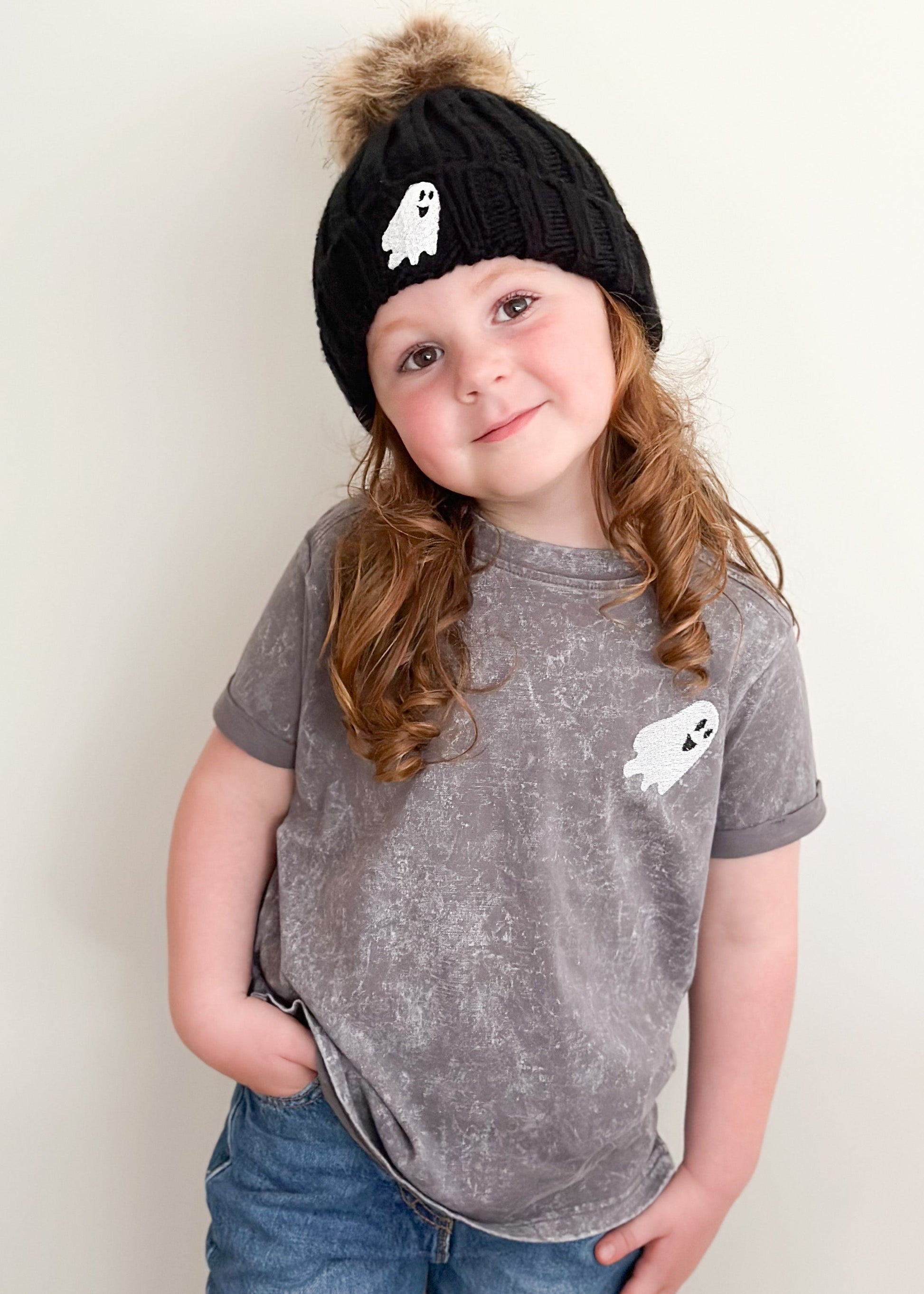 Child wearing a black knit hat with a ghost design and a matching gray t-shirt with a ghost design on a plain background.