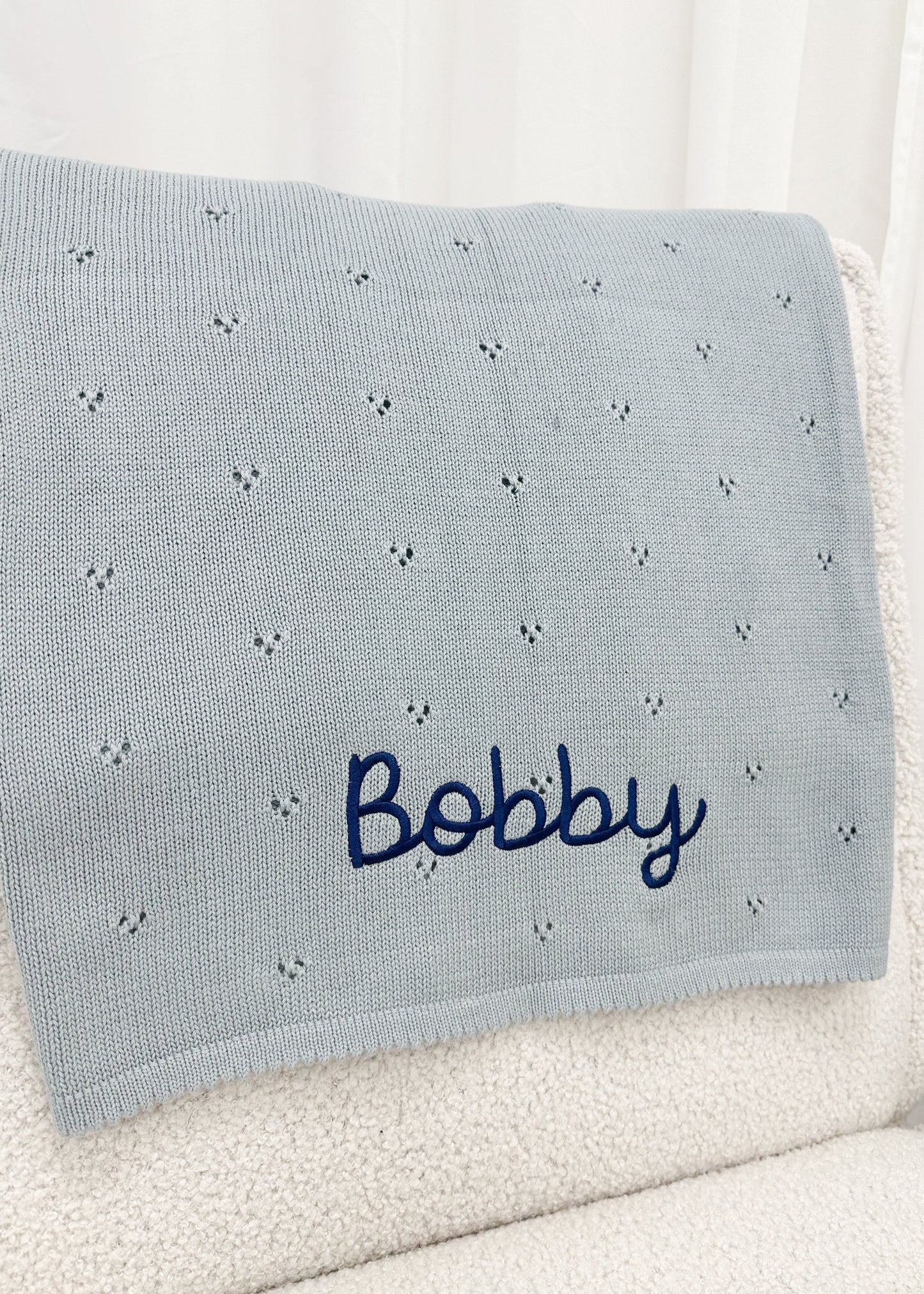 A light blue knitted baby blanket with a pattern of small embroidered faces and the name 'Bobby' embroidered in navy blue script, draped over a white textured chair.