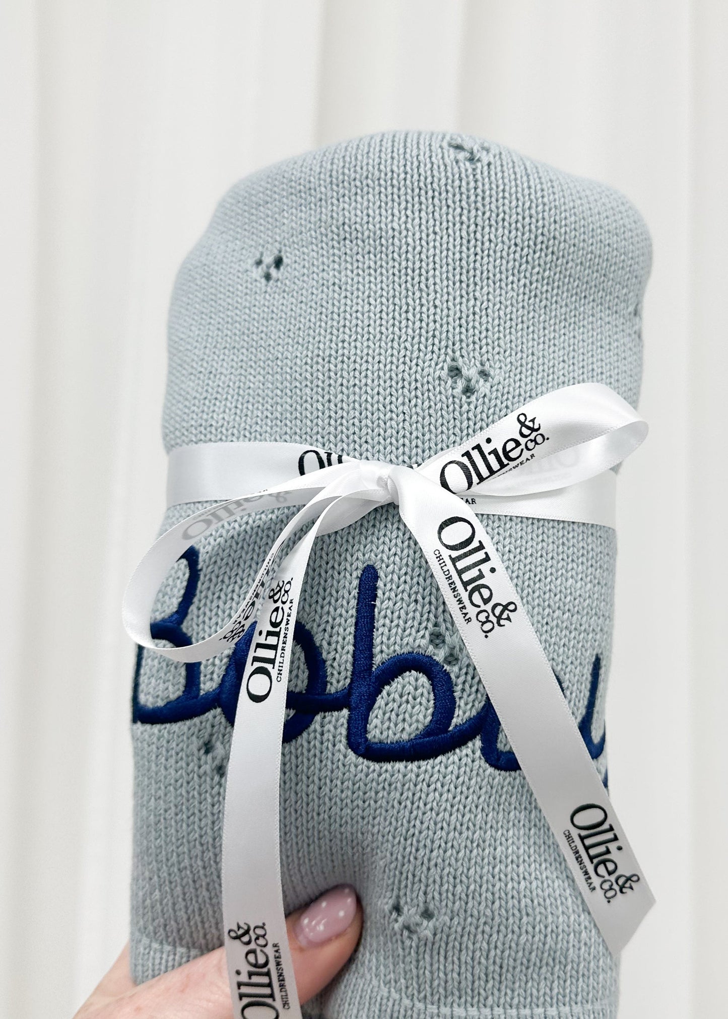 A light blue knit baby blanket with embroidered blue script and small star details, wrapped in a white ribbon printed with the brand name 'Ollie &', held by a hand against a soft white curtain background.