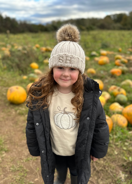 Child in a pumpkin patch wearing a knit hat and coat with a pumpkin design on a t-shirt.