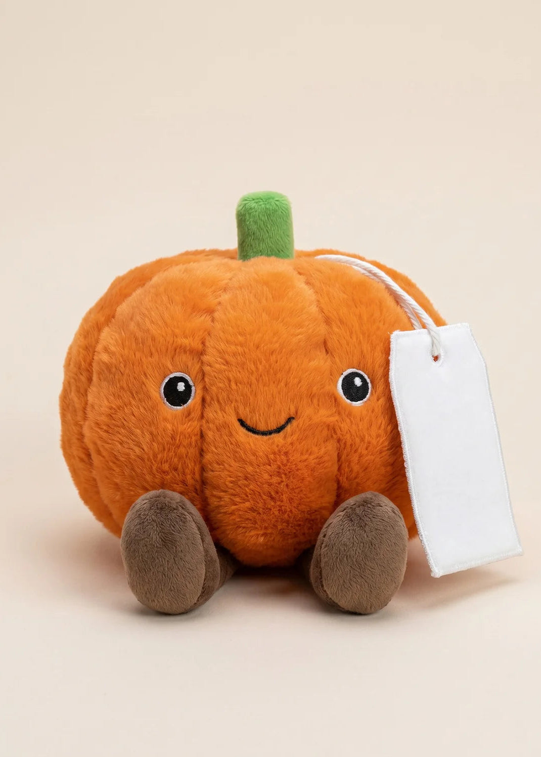 A small orange plush pumpkin toy with a green stem, embroidered smiling face, and brown feet, shown on a plain warm beige background with a blank white tag attached.