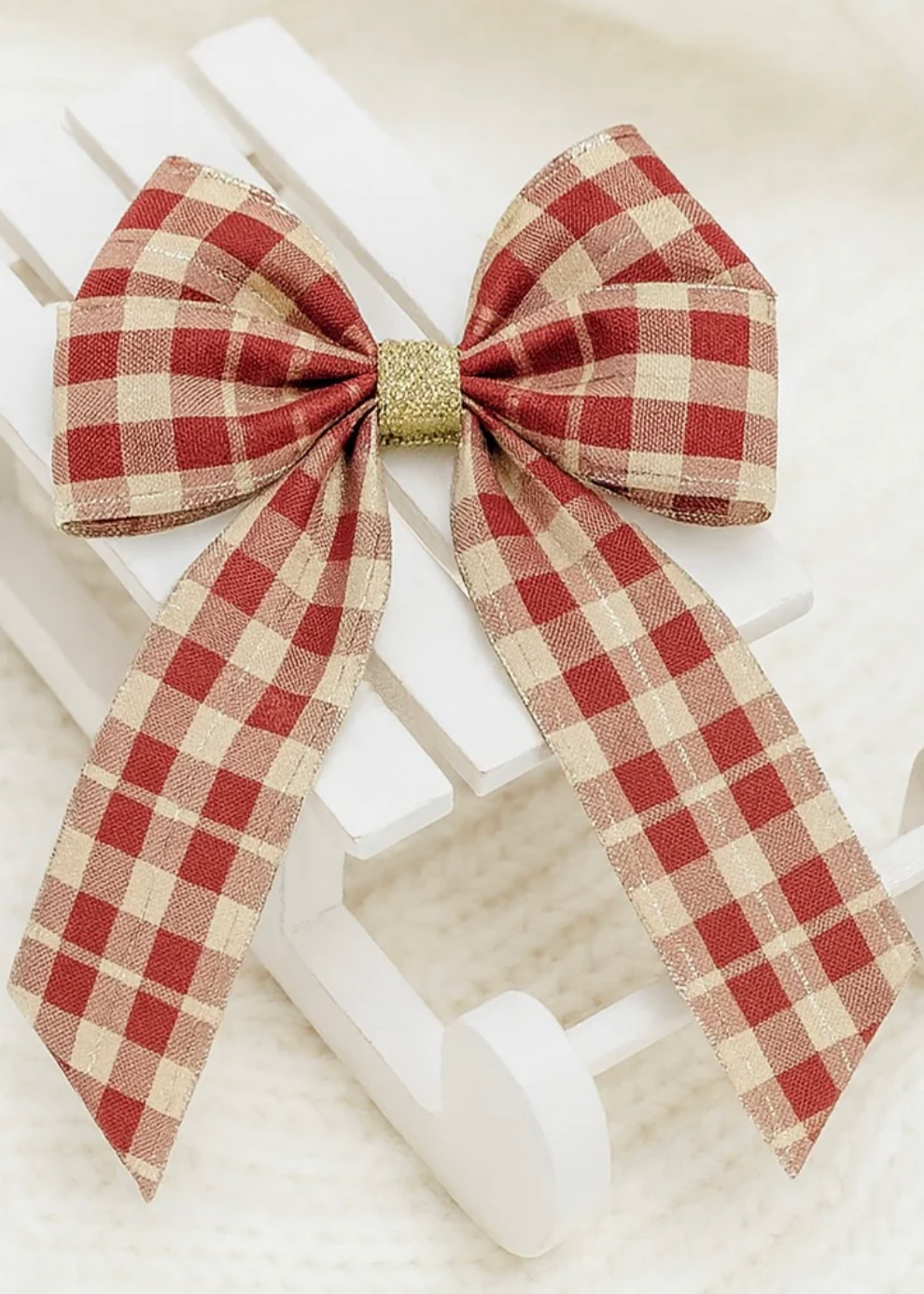 A red and beige gingham check ribbon bow with long tails, secured with a textured gold glitter center, displayed on a white wooden stand against a soft white background.