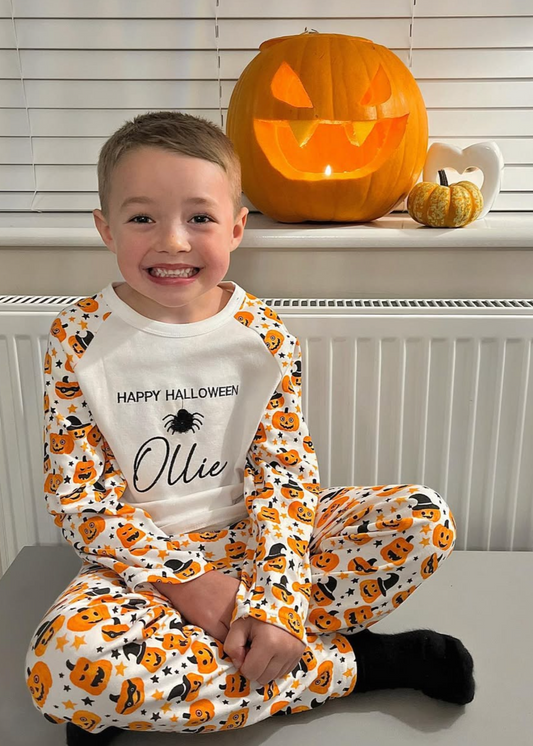 Child wearing Halloween-themed pajamas with a pumpkin design, sitting in front of a carved pumpkin.