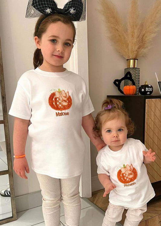 Two children wearing white t-shirts with pumpkin designs and text, standing indoors.