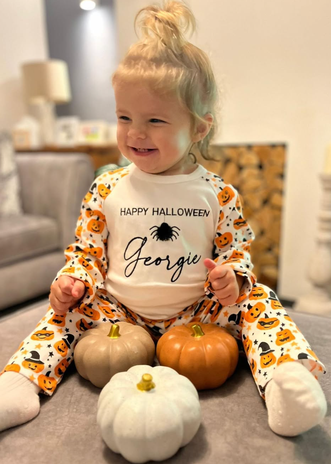 Child wearing a 'Happy Halloween' outfit with pumpkins, sitting on a couch with small pumpkins.