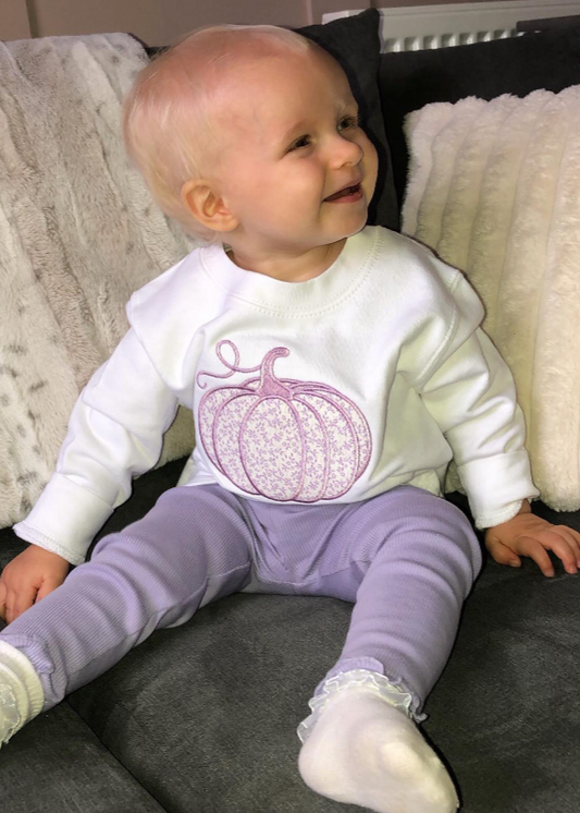 Baby wearing a white sweatshirt with a pink pumpkin design and purple pants sitting on a couch.