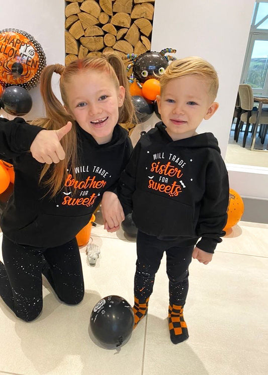 Two children, a boy and a girl, wearing black hoodies with white and orange text, standing in a room with Halloween decorations.